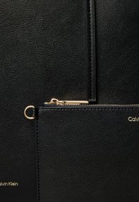 Svart teksturert skinnveske med gullglidelås og ring, som viser en del av Calvin Klein-logoen i gullbokstaver.