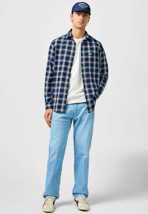 Koszula w kratę flanelowa w kolorze granatowym i jasnoniebieskim, biała koszula z okrągłym dekoltem, jasnoniebieskie dżinsy oraz beżowe sneakersy z czarnymi akcentami. Niebieska czapka z napisem.