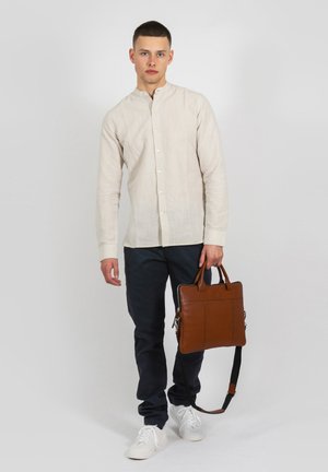 Linen, long-sleeve shirt in beige with a mandarin collar, paired with navy trousers and white sneakers; holding a brown leather bag.