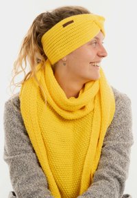 Una donna sorridente con capelli lunghi indossa una fascia in maglia gialla e una sciarpa su un maglione grigio a trama, mentre guarda verso destra.