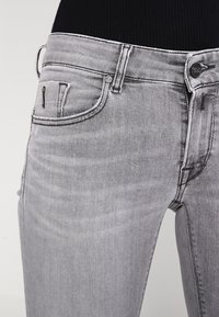 Jean gris de denim con corte Slim, cinco bolsillos y accesorios de metal plateado. Presenta un sutil desvanecimiento y detalles de costura a lo largo de las costuras.
