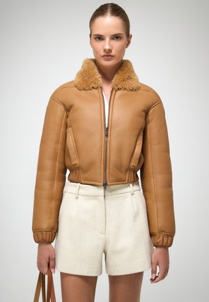 Braune Leder-Bomberjacke mit Fellkragen, gerippten Bündchen und Reißverschluss, kombiniert mit cremefarbenen Shorts, vor einem schlichten Hintergrund.