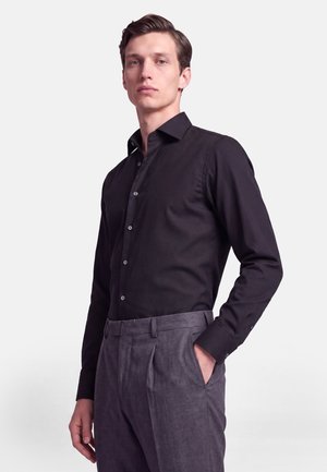 Jeune homme aux cheveux plaqués en arrière, portant une chemise noire boutonnée rentrée dans un pantalon gris taille haute, une main dans la poche.