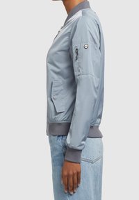 Vue de côté d'une personne portant une veste bomber bleu clair avec des poignets et un col côtelés, associée à un jean bleu clair.