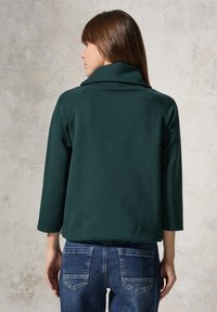 Giacca verde texturizzata con colletto sciallato e maniche 3/4, caratterizzata da un design a fantasia, abbinata a jeans blu, vista da dietro.