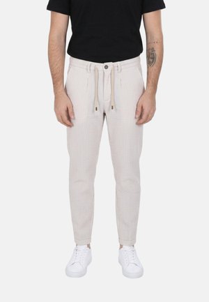Uomo con pantaloni beige e bianchi a righe sottili con coulisse in vita, t-shirt nera, sneakers bianche e tatuaggio sul braccio su sfondo neutro.
