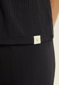 Dark ribbed fabric tank top with a small white label featuring a logo at the hem. The texture is smooth with vertical lines.