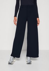 Vida, marinblå byxor i slät tyg med en skräddarsydd midjeband. Kombinerade med silver- och grå sneakers, vilket visar en avslappnad stil.