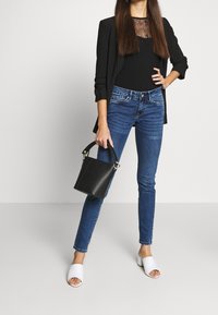 Kvinde iført sort blazer og blonde top, blå skinny jeans, hvide åbne hæle, holder en sort bøtte-handbag, står opad en ensfarvet baggrund.