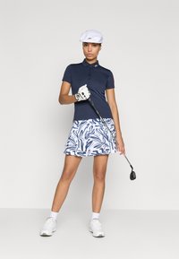 Tumši zils polo krekls ar pogāmo apkakli, ar rakstiem balts un zils šorti, balti golfa apavi un balta cepure. Tur rokās golfa nūju.