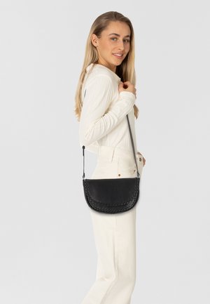 Schwarze Ledertasche zum Umhängen mit glatter Textur und geflochtenem Akzent entlang der Kante, getragen über einem weißen Langarmtop und hellen Hosen.