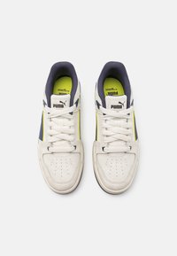Un paio di sneakers Puma bianche con dettagli neri e gialli, lacci bianchi e solette giallo neon viste dall'alto su uno sfondo bianco.