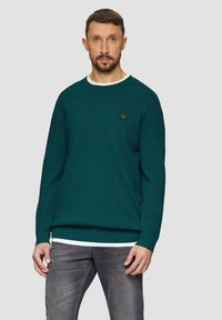 Dunkelblaugrüner Strickpullover mit Rundhalsausschnitt, langen Ärmeln und einer weißen Innenschicht, die hervorblitzt. Mit einem kleinen Logo-Patch auf der Brust.