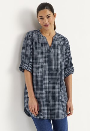 Kvinde iført en mørkeblå ternet skjorte med opkrogede ærmer og blå jeans, smilende, stående mod en hvid væg.