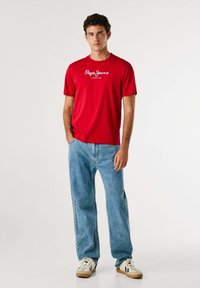 Rdeča kratka majica s kratkimi rokavi z belim napisom "Pepe Jeans London", v kombinaciji z svetlo modrimi kavbojkami in bež supergami z črnimi detajli.