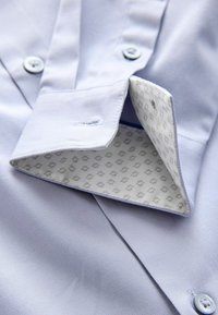 Camisa azul claro de manga larga con una textura suave, que cuenta con cierre de botones y un cuello interior con patrón en blanco con diseños grises.