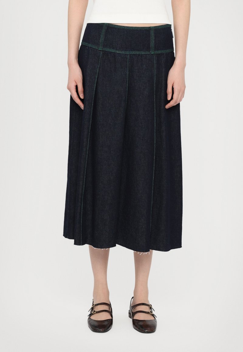 Mørk denim midiskjørt med grønn søm, med flat midjebånd og pregede detaljer. Kombinert med brune flate sko.