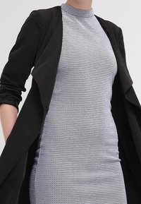 Czarna zewnętrzna kurtka nałożona na dopasowaną szaro-siatkową sukienkę z wysokim dekoltem, z teksturowanymi wzorami i gładkim wykończeniem.