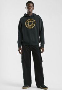 Schwarzer Hoodie mit einem strukturierten Golddruck, der "QUITE SENSE" liest, kombiniert mit weiten schwarzen Cargo-Hosen mit Taschen und lockerer Passform.