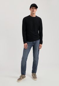 Schwarzer Langarmpullover, lässige blaue Jeans, beige und graue Sneakers, schwarze Mütze, stehend vor einem neutralen Hintergrund, lockere Passform, keine sichtbaren Muster.