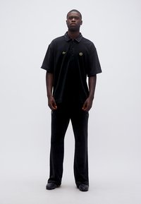 Mann steht mit Blick nach vorne, trägt ein schwarzes Adidas-Poloshirt und lockere schwarze Hosen vor einem einfarbigen hellen Hintergrund.