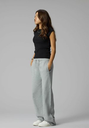 Jeune femme debout en pantalon de survêtement gris, haut sans manches noir, baskets blanches, regardant à gauche avec les mains dans les poches sur fond gris uni.