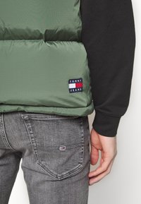 Zelená prošívaná vesta s vodorovným prošitím a nášivkou s logem Tommy Jeans. Kombinována se šedými džínami, které na kapse mají detail s logem.