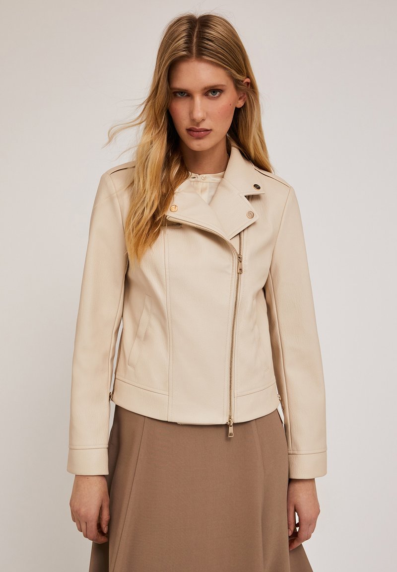 Giacca motociclista in similpelle beige con zip diagonale, bottoni decorativi e una finitura testurizzata sottile. Abbinata a una gonna beige sottostante.