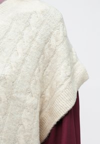 Kremno pletivo s kabli, pončo z ribastim robom. Mehak material in ohlapen kroj, položen preko temno burgundskega oblačila.