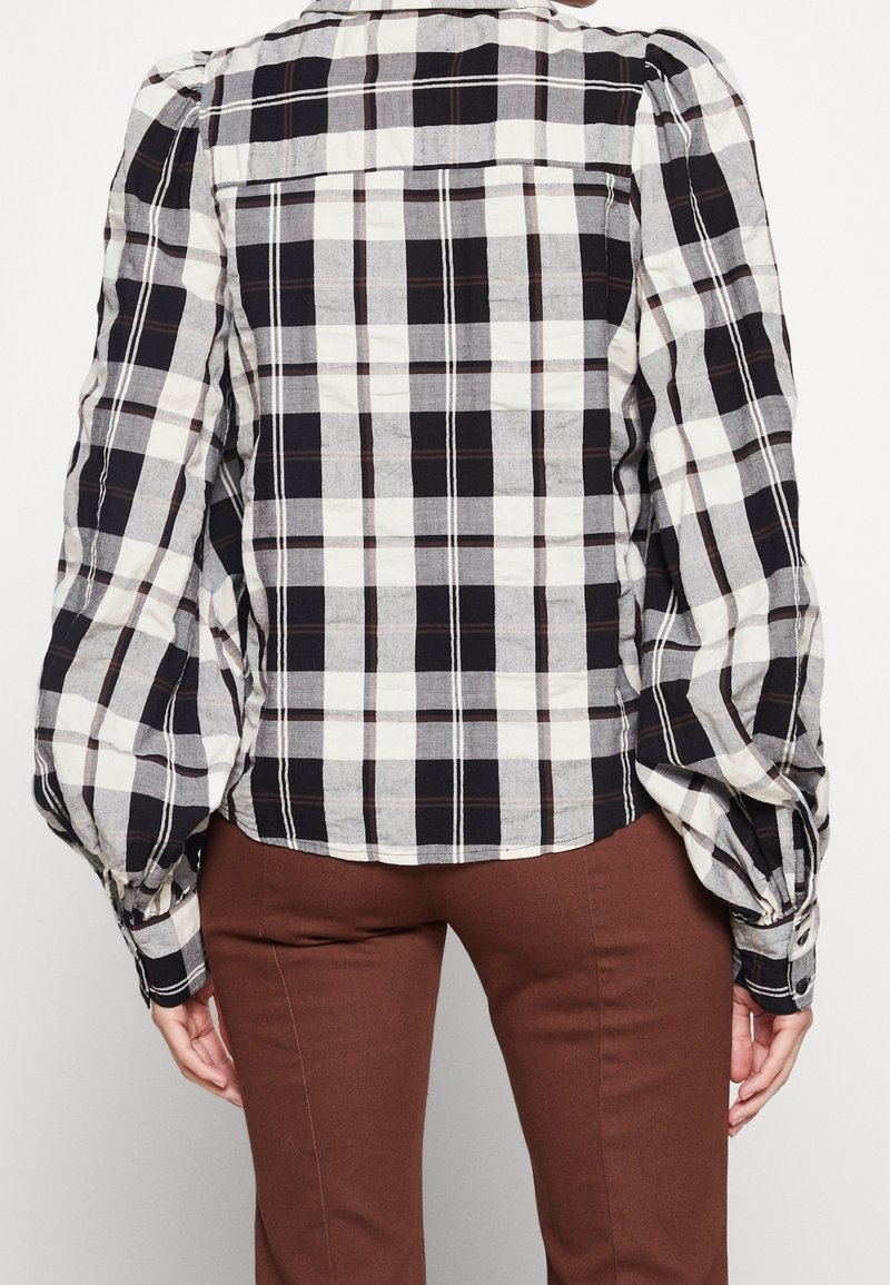 Chemise à carreaux noir et blanc avec des manches bouffantes, fermeture à boutons et ourlet arrondi, associée à un pantalon marron pour un look superposé.
