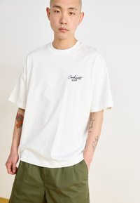 Camiseta blanca de algodón con cuello redondo, mangas cortas y un logo negro bordado en el lado izquierdo del pecho. Combinada con pantalones cortos de color verde olivo.