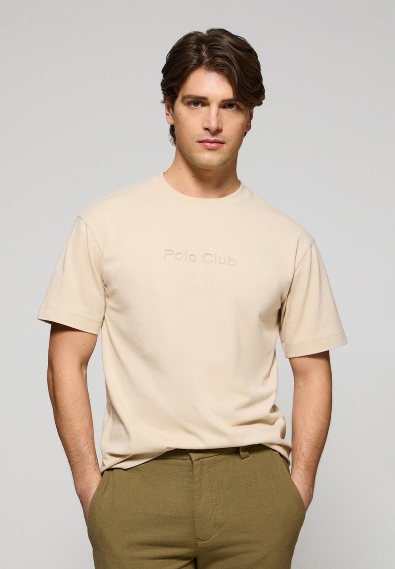 Beige bomullströja med korta ärmar, prydd med präglad "Polo Club"-text på framsidan. Slät textur, i kombination med olivgröna byxor.