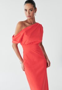 Model u haljini s jednim ramenom u crvenoj boji stoji pred bijelom pozadinom, prikazujući samopouzdanje i eleganciju. Dodatno se uređuje naušnicama i narukvicom.