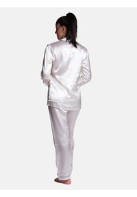 Pigiama in raso bianco composto da una maglia a maniche lunghe e pantaloni dritti, caratterizzati da una texture liscia e da piping a contrasto lungo i bordi.