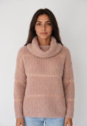 Femme aux longs cheveux foncés portant un pull à col roulé rose pelucheux et un jean bleu clair, debout sur un fond uni clair.