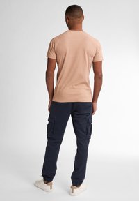 Hnědé tričko s krátkým rukávem, kombinované s námořnickými cargo kalhotami s mnoha kapsami, elastickými manžetami a béžovými teniskami. Jednoduché pozadí.