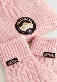 Roze gebreide muts en handschoenen set, met kabelbrei textuur, geribde manchetten en een ronde logo patch met "SUPERDRY" merknaam.