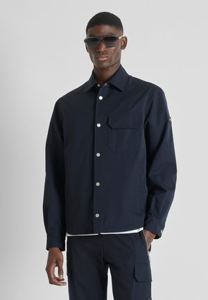 Man die een donkerblauwe jas met knopen en bijpassende cargobroek draagt, met een zonnebril op, staand tegen een eenvoudige lichte achtergrond.