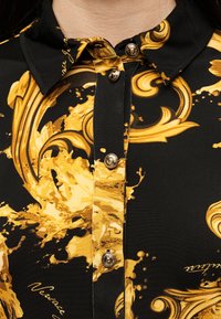 Μαύρο πουκάμισο με χρυσά μπαρόκ στροβιλιστικά σχέδια, χρυσά κουμπιά με περίτεχνα σχέδια και διπλωμένο μαύρο γιακά.