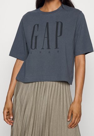 Szara, krótka koszulka z logo "GAP" w czerni, z napisem "1969" poniżej, wykonana z bawełny, zestawiona z plisowaną beżową spódnicą.