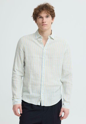 Ung mand med krøllet hår iført en lysefarvet, vertikalt striped skjorte med knapper og mørke bukser, stående foran en ensfarvet baggrund.