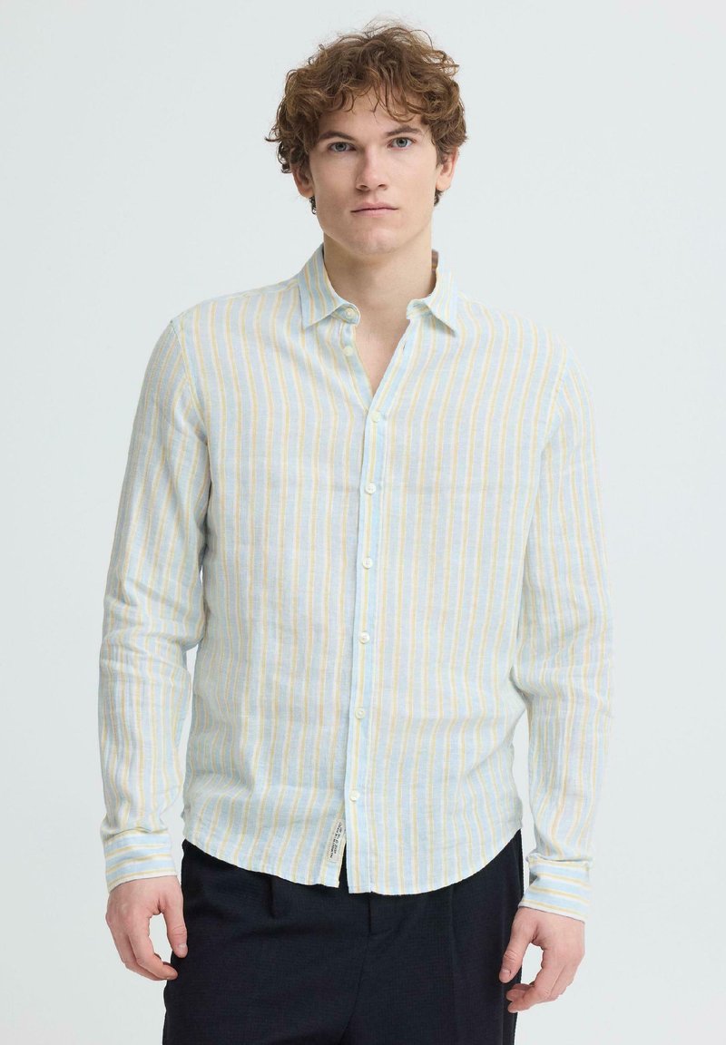 Jeune homme aux cheveux bouclés portant une chemise à boutons à rayures verticales claires et un pantalon foncé, debout devant un fond uni.