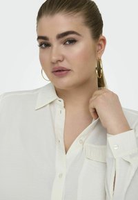 Cămașă albă cu guler, dotată cu un buzunar, având un material texturat. Modelul are o coadă de cal și poartă cercei mari din aur.