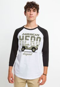 Hvid t-shirt med sorte 3/4 ærmer, der har grafisk tekst "American Hero" og en illustration af en grøn 1944 Willys MB Jeep.