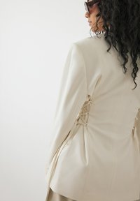 Blazer entallado beige con detalles de cordones en la parte trasera, hombros estructurados y textura suave. Corte oversized con mangas largas.
