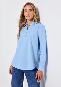 Camicia azzurro chiaro con colletto, apertura con bottoni, maniche lunghe e orlo leggermente curvato, realizzata in tessuto morbido. Indossata con jeans in denim.