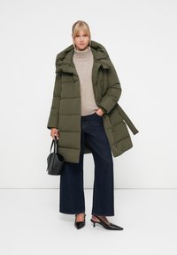 Olive green puffer coat with a hood, side pockets, and a cinched waist. Worn over a beige turtleneck and paired with wide-leg jeans and black heels.
