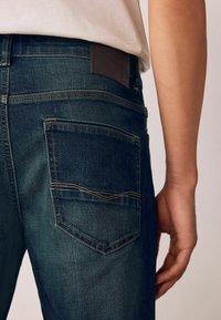 Tumed sinised denim teksad, millel on nähtav tagatasku, kontrastne õmblus ja nahk silt vööl. Sile tekstuur ja kitsas lõige.