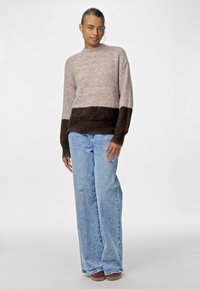 Strickpullover in Beige und Dunkelbraun, mit Rundhalsausschnitt und langen Ärmeln. Kombiniert mit hellblauen weit geschnittenen Jeans und lässigen Schuhen.