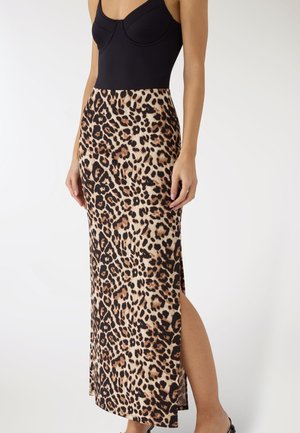 Kobieta ubrana w czarny top bez rękawów oraz midi spódnicę w panterkę z bocznym rozcięciem, stojąca na tle jednolitego białego tła.